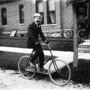 A man poses on a bicycle in Denver, Colorado; his outfit includes a straw hat. A brick and stone building has a covered porch.