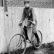 A boy poses on a bicycle by wooden steps in Denver, Colorado; his outfit includes a messenger hat, a knit sweater, and a ribbon or medal.