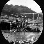 Seven African American campers from Camp Nizhoni in Lincoln Hills (Gilpin County), Colorado either stand in South Boulder Creek or sit on a wooden bridge. They wear swimsuits and swim caps.