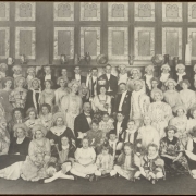 Men, women and children pose in costume for a group portrait at probably the Albany Hotel parlor on 17th (Seventeenth) and Stout Streets in downtown Denver, Colorado. Women and men wear seventeenth or eighteenth century court costume and wigs or tuxedos and pose in front of glass tile or stained glass windows. The (probably) Apple Queen wears a crown and holds a staff with ribbons.