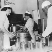 Women and an African American man can food probably for school lunch programs in Denver, Colorado. Shows cans, large pots and pressure cookers.
