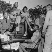 African American soldiers in uniform and women laugh as they stand near, or sit on, a piano at a W.P.A. armed forces center in Denver, Colorado. The women wear dresses, one has on strap heeled shoes.
