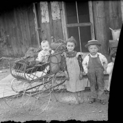 Outdoor portrait of children, they include Lydia Lillybridge (Medin), Montana Lillybridge (Ericson), and a baby in a carriage near a house in Denver, Colorado. The boy wears overalls and a straw hat.