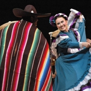 View of unidentified Mexican folk dancers at a Cinco de Mayo celebration in Denver, Colorado. A woman in a blue costume decorated with purple and white frills dances with a man wearing a Mexican serape and a sombrero. The woman has ribbons and a bow in her hair.