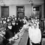 Interior view of a soup kitchen for the unemployed in Denver, Colorado; African American (Black) and white men and boys sit or stand at tables with bowls of soup and bread. Shows a coffee urn and cafeteria employees.