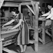 A Hispanic American man and woman work on traditional wooden treadle looms probably in Southern Colorado. The woman works on a Rio Grande style weaving.