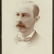 Studio portrait of a man with a bow tie, Autolycus style moustache and beard patch, and thin hair.