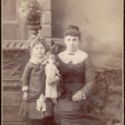 Studio portrait of a woman (seated), a girl( standing) in front of a prop balustrade and potted plant. The girl holds a doll and wears a pleated skirt and a long top with a lace collar and a bow at the waist, her hair is in ringlets and she has a cord around her neck with a pendant. The woman wears a long dress with a crocheted collar and a pin, Her hair is in a bun.