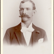 Studio portrait of a man with a waxed handlebar moustache, bow tie, and boutonniere.