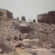 View of a Manitou and Pike's Peak Railway locomotive and passenger car at the summit of Pikes Peak in El Paso, County, Colorado. People wait in a boulder field outside the stone Army Signal Station; a pack burro is in the foreground.