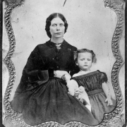 A studio portrait of Elizabeth Minerva Byers (Mrs. William N. Byers) and her daughter Mary (later Mary Byers Robinson). Mrs. Byers wear a dark dress and a brooch, her daughter wears a checked off the shoulder dress.