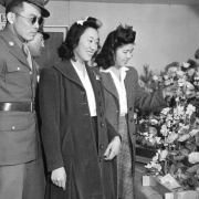 Two Japanese soldiers in uniform pose with their girlfriends by a flower exhibit, at the Arts and Crafts Festival, Granada Relocation Center, Camp Amache, Prowers County, southeastern Colorado. The women wear long wool coats, white blouses, skirts, and ribbons in their curled hair. One holds an artificial flower in the display. The soldiers wear glasses and sunglasses.