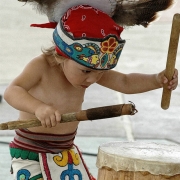 Three year old Chico Stange, a member of Azteca Art Colorado, plays a carved wooden drum in Denver, Colorado. He holds two carved, wooden drum sticks. He wears a feather headdress decorated with beadwork, and a fabric loincloth and apron decorated with Aztec patterns highlighted with glitter.