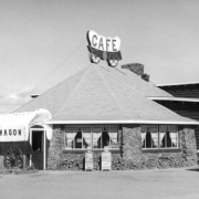 Exterior of the Chuck Wagon Cafe and Cocktail Lounge, Granby, Grand County, Colorado, shows the stone restaurant with pyramidal roof, covered wagon entrance, and painted log back addition. A neon covered wagon  sign tops the roof. Rocky Mountain News and Denver Post newspaper boxes stand at the entrance. Signs in curtained windows read: "Help Wanted," "Come In We're, Open," and "Yes...We Are Open."