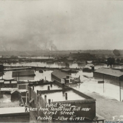 View of the flooded Arkansas River in Pueblo (Pueblo County), Colorado. People stand and look at the destruction from railroad tracks in Goat Hill (Tenderfoot Hill) in northeast Pueblo. Shows a railroad bridge, buildings submerged in water, probably the Nuckolls Packing Company building, and smokestacks in the distance.