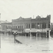 View of flood water and submerged commercial buildings at the intersection of Main Street and Union Avenue in Pueblo (Pueblo County), Colorado. Shows debris on a fallen telephone wire, and signs on businesses that read: "Stark Clothing Co.," "The Wonder Men's Store," "United Shoe Stores Co.," "Central Studio," and "Central Market."