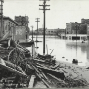 People pose on a freight car and on a pile of debris beside flood water from the Arkansas River on 1st (First) Street in Pueblo (Pueblo County), Colorado. Shows a pile of lumber, a wagon wheel, and flood water on First Street. Brick buildings read: "Haver's Land Office," "T.G. McCarthy and Co. Undertakers," and "Auto Parts & Rubber House, United States Tires, Sales & Service Depot."