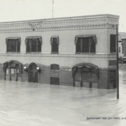 View of a two-story brick building with a flat roof and cloth awnings identified as the "Old White Triangle Block" on Union Avenue and Main Street in Pueblo (Pueblo County), Colorado. Flood water from the Arkansas River surrounds commercial businesses. Shows submerged doorways, windows, street lights, telephone poles, and piles of debris.