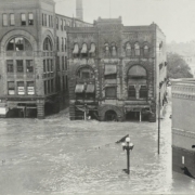 View of Arkansas River flood water at the intersection of Main Street and Union Avenue in Pueblo (Pueblo County), Colorado. Flood water is half way up the first story of stone commercial buildings with arched doorways and windows, and flat roofs. Flood debris is on power lines in the street.