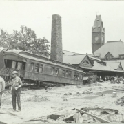 Men stand outside the Union Depot in Pueblo (Pueblo County), Colorado. One man wears a badge. Shows water, mud and debris on the ground, and a flood damaged passenger car wrecked beside the stone depot. Probably a cleaning crew works on mud covered tracks nearby.