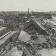 View of wrecked Denver and Rio Grande Western freight cars and frame buildings after the Arkansas River flood in Pueblo (Pueblo County), Colorado. Piles of lumber and debris, mud and silt are in the Walker rail yard. A smokestack is beside a building that reads: "This Property for Sale or Rent, apply to H.C. Borndruck, Phone 120."