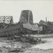 View of an Atchison, Topeka & Santa Fe railroad bridge and a wrecked Denver & Rio Grande Western railroad bridge with piles of wreckage from the Arkansas River flood in Pueblo (Pueblo County), Colorado. Men stand and pose on piles of debris and rubble from flooded buildings and rail yards.