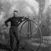 Studio portrait of a man, possibly W.F. Blake. He leans on a high bicycle and wears special riding pants, turtle neck shirt, and cap. He has a handlebar moustache.