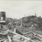 Men work to clear rubble piled along the flooded Arkansas River in Pueblo (Pueblo County), Colorado. Freight cars, a locomotive, hay bales, tracks and lumber are scattered near Atchison Topeka & Santa Fe and Denver & Rio Grande Western railroad bridges.