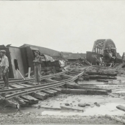 Men in overalls or work clothes and hats stand on washed out railroad tracks beside piles of rubble from the Arkansas River flood in Pueblo (Pueblo County), Colorado. Atchison Topeka & Santa Fe and Denver & Rio Grande Western railroad bridges are in the distance. Shows wrecked and overturned locomotives and railroad cars.