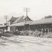 View of the 8th (Eighth) Street Depot beside Denver and Rio Grande Western Railroad tracks in Pueblo (Pueblo County), Colorado. Shows mud covered tracks and debris from flooded Fountain Creek washed up beside the covered passenger platform. The frame depot building has a hipped roof, half-timbered dormer windows and a sign that reads: "Eighth Street Pueblo."