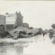 People walk on 2nd (Second) Street in front of damaged commercial buildings and piles of debris from the Arkansas River flood in Pueblo (Pueblo County), Colorado. Men pose on wreckage from a collapsed roof beside a damaged brick building with a decorative parapet. Shows cars, water and mud in the street.