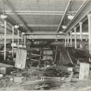 Interior view of White and Davis clothing store at 301 N. Main Street after the Arkansas River flood in Pueblo (Pueblo County), Colorado. Shows wrecked furniture, crates, mud and debris in the aisle. Signs for "Boys Dept." and "Collars" are posted on pipe columns with Corinthian capitals. The store has a pressed metal ceiling and overhead lamps with glass globes.