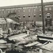 Debris from the Arkansas River flood at 2nd (Second) Street and Grand Avenue in Pueblo (Pueblo County), Colorado. Shows mud, timber, portions of ceiling molding, and a door near the wrecked, frame "Mammoth Corral" building. The three story brick "Colorado Bedding Co." building shows paint chipped off at the high water mark above first story windows.