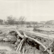 A man in overalls and a fedora stands beside a debris covered Atchison, Topeka and Santa Fe Railroad bridge and a mud covered Denver and Rio Grande Western bridge in probably Pueblo County, Colorado. Tree branches and mud are washed up against the bridge and over the sets of railroad tracks after a flood. Shows an eroded river bed of probably the Arkansas River.