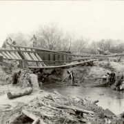 Men work to clear debris from a flood under washed out Denver and Rio Grande Western Railroad bridge number "126-B" in probably Pueblo County, Colorado. Shows tree branches and mud in an eroded creek bed, and unsupported tracks over the creek.