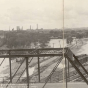 Panoramic view of flood damage from the Arkansas River flood in Pueblo (Pueblo County), Colorado. Shows washed out Denver and Rio Grande Western Railroad tracks and the rail yard near the Union Depot, mud and debris on the ground and smokestacks in the distance. Men stand near a car on the West 4th (Fourth) Street bridge.