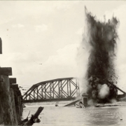 View of a dynamite explosion to dislodge a Denver and Rio Grande Western Railroad bridge from flood debris in the Arkansas River in Pueblo (Pueblo County), Colorado. Shows mud and sand in the air, part of a D. & R.G. bridge submerged in the river and an Atchison, Topeka and Santa Fe Railroad bridge nearby.