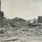 Piles of debris from the Arkansas River flood are in the streets around the stone Atchison Topeka and Santa Fe Railroad depot in Pueblo (Pueblo County), Colorado. Lumber, metal, mud and silt cover the streets near brick commercial buildings.