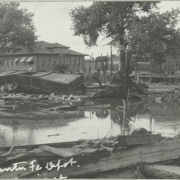 View of overturned freight cars and wreckage from the Arkansas River flood on Union Avenue in front of the stone Atchison, Topeka and Santa Fe Railroad depot in Pueblo (Pueblo County), Colorado. Shows standing water around trees and piles of debris.