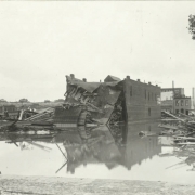 View of standing water piles of lumber and debris on Main Street and Hobson Street after the Arkansas River flood in Pueblo (Pueblo County), Colorado. Shows the collapsed wall of the Ford Exchange building.