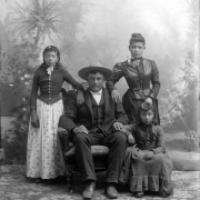Studio portrait of possibly the Horea Baskes (Vasquez) family (Hispanic). The man wears a dark wool suit, a wide brimmed hat, and sits in an upholstered chair. His wife stands at his side. She wears a two-piece dark colored dress with a fitted bodice that buttons up the front. Her hair is pulled to a bun on the top of her head. A young girl wears a dark colored wool bodice with a light colored print cotton skirt and gloves. Another girl wears a gray wool dress with a ruffle.
