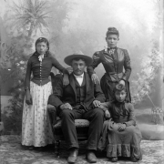 Studio portrait of possibly the Horea Baskes (Vasquez) family (Hispanic). The man wears a dark wool suit, a wide brimmed hat, and sits in an upholstered chair. His wife stands at his side. She wears a two-piece dark colored dress with a fitted bodice that buttons up the front. Her hair is pulled to a bun on the top of her head. A young girl wears a dark colored wool bodice with a light colored print cotton skirt and gloves. Another girl wears a gray wool dress with a ruffle.