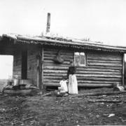 A woman holds a child's hand at the side of a log cabin, Goldfield, Teller County, Colorado. The front gable house is constructed of logs and hewn lumber and has one glass pane window, a mud and sod roof with a stove pipe. Several wooden boxes and crates are piled in front of the cabin and a full sack is hung on the side. A man in a horse-drawn wagon shows in distant far left.