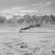 A Denver and Rio Grande Western narrow gauge train pulled by locomotive 480 heads northwest and vents smoke near Villa Grove (Saguache County), Colorado. Clouds and snow top the Sangre De Cristo range.