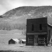 View of a dilapidated two-story wood frame false front with a corrugated metal gable roof, dentiled cornice, two upper-story windows, remains of an outside side stairway and a single door entry flanked by windows in Gothic, Gunnison County, Colorado. A faded sign reads: Lee's Tavern; two small gabled sheds show in background. Gothic was a mining boom town from 1879 to 1884; bought, in 1928, by the Rocky Mountain Biological Laboratory for plant and animal research.