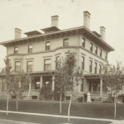 John Kernan Mullen and Catherine Smith Mullen pose outside their three-story house at 896 Pennsylvania Street in the Capitol Hill neighborhood of Denver, Colorado. The brick house has a rusticated stone foundation, covered porches with overhanging, bracketed eaves, columns and dentils, second-story bay windows, roof dormers and chimneys. Mr. and Mrs. Mullen stand on a manicured lawn near trees, bushes and a lawn sprinkler.