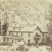 Men and women pose in front or on the second-story balcony of the Barton House hotel in Georgetown (Clear Creek County), Colorado. Men sit on a horse-drawn stagecoach. The frame hotel has a hipped roof with dormer windows, brackets and overhanging eaves. Shows snow on mountains and near tree stumps.