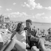 A Tenth Mountain Division soldier enjoys the beach at Trieste with a woman. Both wear bathing costumes and pose for the camera. Behind them the beach is filled with other sun worshipers.