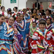Girls, members of Jeanette Trujillo-Lucero's Fiesta Colorado Dance Company pose near Civic Center Park during Cinco de Mayo celebrations in Denver, Colorado. The girls wear traditional Mexican style dance costumes in bright colors decorated with lace and ribbon. They all have their hair pulled up with flower decorations.