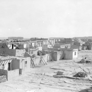 A row of small buildings of the Laguna Pueblo in New Mexico is shown from the back. Wooden ladders lead to the upper levels of the structures. The windows and doors are framed with wood. The surface of the ground is composed of large slabs of rock. Small mounds, probably ovens, stand a few feet behind the buildings. Additional mesas, including the "Enchanted Mesa," are in the distance.